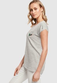 T-shirt gris à manches courtes présentant un design de cœur et de chaussure brodé sur la poitrine, porté avec une coupe décontractée sur un pantalon blanc.
