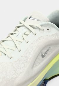 Nike obuća s teksturiranim svijetlosivim gornjim dijelom, bijelim detaljima i žuto-zelenom potplatu. Ima srebrni logo i funkcionalni sustav vezivanja.