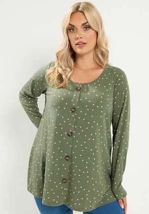 Blondynka ubrana w oliwkowy top z długim rękawem z białymi kropkami i brązowymi guzikami, w połączeniu z niebieskimi jeansami.
