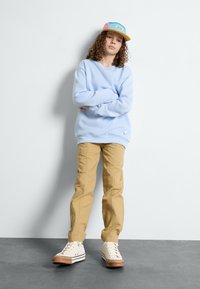Hellblaues Fleece-Sweatshirt, khakifarbene Cargo-Hosen, weiße Sneakers mit braunen Sohlen und eine mehrfarbige Kappe mit einem Logo auf der Vorderseite.
