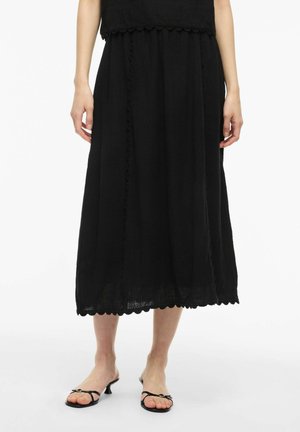 Person iført et sort midi-nederdel med bølgede kanter og sorte hælsandaler med tynde stropper, stående på en hvid baggrund.