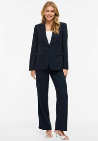 Femme souriante aux longs cheveux blonds portant un costume sombre à fines rayures, un haut blanc et des sandales à talons ouvertes blanches, debout devant un fond blanc.