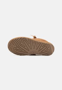 Sandale beige avec une tige texturée, dotée d'une large sangle sur l'avant-pied et d'une semelle en caoutchouc plate et à motif. Le logo de la marque est visible sur la semelle.