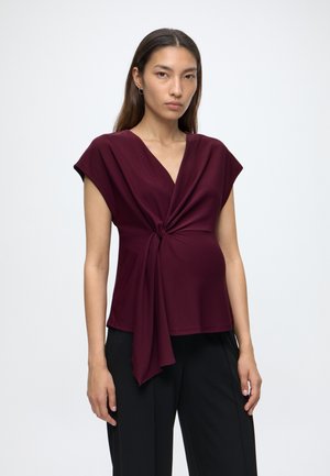 Femme aux longs cheveux foncés portant un haut bordeaux sans manches avec un nœud sur le devant et un pantalon noir sur un fond uni.