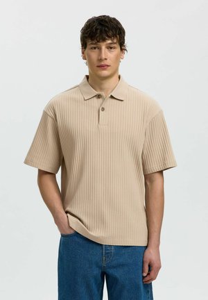 Junger Mann mit kurzen dunklen Haaren trägt ein beiges geripptes kurzärmeliges Polohemd und blaue Jeans, steht mit einer Hand in der Tasche.