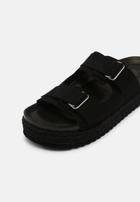 Svart platt slid sandal med två justerbara remmar och silvriga spännen, strukturerad flätad sula och vadderad fotbädd på vit bakgrund.