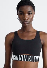 Kvinne med kort hår iført svart Calvin Klein trenings-BH med hvit logo på elastikken, vendt mot kameraet mot en ensfarget bakgrunn.