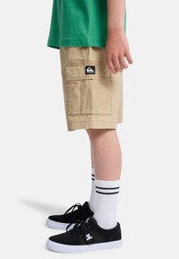 Beige Cargo-Shorts mit Klappentasche und Quiksilver-Logo, getragen mit weißen Socken und schwarzen DC-Sneakern an einer stehenden Person.