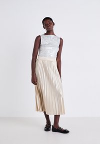 Sleeveless, shiny silver top paired with a pleated beige skirt. Model wears black shoes with decorative hardware details. Plain background.