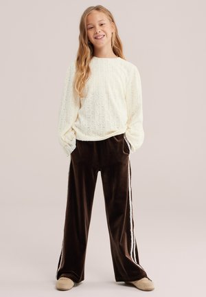 Fille souriante aux longs cheveux blonds portant un pull blanc texturé, un pantalon large foncé avec des bandes latérales et des chaussures beiges, debout les mains dans les poches.