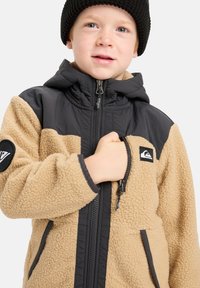 Chaqueta de forro polar marrón con acentos de nailon negro, cierre frontal con cremallera y bolsillos, capucha y parche con logotipo en el pecho. Diseño texturizado y cálido.