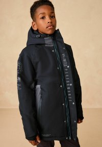 Schwarze wasserdichte Jacke mit grauen Akzenten, ausgestattet mit einem hohen Kragen, Reißverschluss, mehreren Knöpfen und bedrucktem Text auf der Vorderseite.