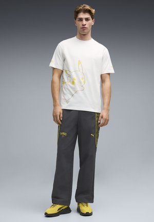 Jeune homme portant un t-shirt blanc imprimé Pikachu, un pantalon foncé avec les logos jaunes Puma et Pokémon, et des baskets jaunes, debout sur un fond gris.