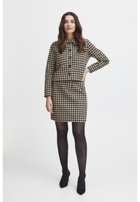 Sukienka w wzór houndstooth w beżu i czerni, z kołnierzykiem, długimi rękawami i dopasowaną spódnicą. W zestawie z czarnymi rajstopami i szpilkami.