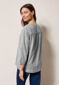 Chemisier rayé à boutons en gris et blanc, avec un col rond, un dos froncé et des poignets élastiques. Associé à un jean bleu.