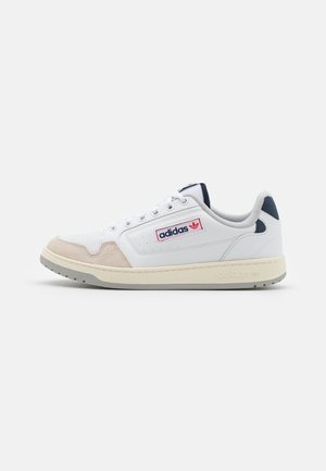 Zapatilla blanca de Adidas con acentos en ante beige, suela gris, lengüeta de talón azul marino y parche con el logo de Adidas en el lateral, mostrada de perfil sobre un fondo liso.
