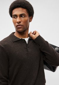 Σκούρο καφέ πλεκτό πόλο μπλούζα με ριγέ γιακά και μανσέτες, με δύο κουμπιά στο λαιμό. Το μοντέλο κρατά μια μαύρη υφασμάτινη τσάντα.