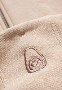 Tejido beige con una textura suave que presenta un logo bordado en espiral marrón. Incluye una cremallera a juego para el cierre.