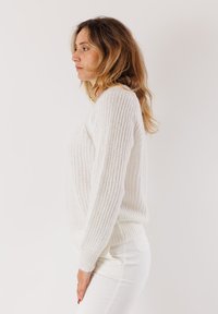 Donna con capelli castano chiaro mossi, che indossa un maglione bianco di maglia e pantaloni bianchi, rivolta a sinistra contro uno sfondo chiaro uniforme.