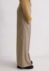 Persoon in beige geruite wijde broek, een bruine trui en donkerbruine schoenen, die zijwaarts tegen een effen witte muur staat.