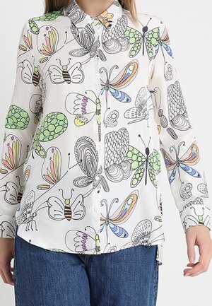 Chemise blanche à manches longues boutonnée avec des motifs colorés et abstraits de papillons et d'insectes, portée avec un jean bleu.