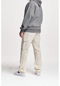 Persoon in lichtbeige cargobroek, grijze hoodie en witte sneakers met zwarte swoosh, staand tegen een eenvoudige witte achtergrond.