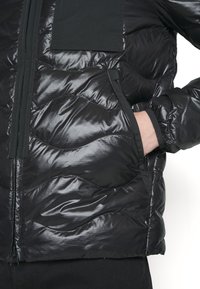 Veste en duvet noire avec une texture brillante et matelassée, dotée d'une fermeture éclair à l'avant, de poches latérales et de poignets élastiques. Comprend un écusson noir uni sur la poitrine.