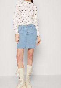 Frau trägt ein weißes Langarmoberteil mit roten Herzen, einen hellblauen Denim-Rock und cremefarbene Schnürstiefel und steht vor einem schlichten Hintergrund.