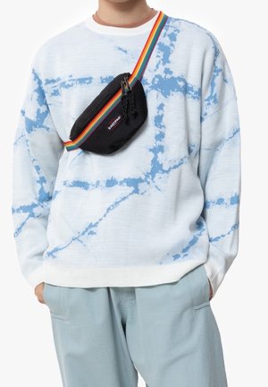 Persoon draagt een lichtblauw tie-dye trui en broek met een zwarte Eastpak crossbodytas met een regenboogband over de borst.