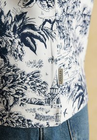 Nahaufnahme eines weißen Gewebes mit dunkelblauem Toile-Druck, der Bäume, eine Kirche und eine Landschaft zeigt, mit angenähtem Etikett "Joules", kombiniert mit blauer Jeans.