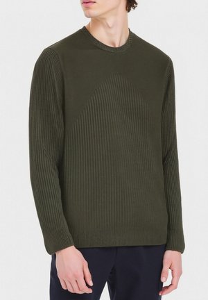 Uomo che indossa un maglione a maniche lunghe a coste di colore verde oliva con un motivo texturizzato curvo nella parte anteriore, abbinato a pantaloni blu scuro, mentre è in piedi davanti a uno sfondo bianco.