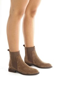 Botines cortos de ante marrón con textura suave, paneles laterales elásticos y un tacón bajo apilado. El diseño presenta una punta redondeada.