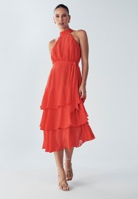 Rode halternekjurk met gelaagde ruches, lichte stof, ceintuur in de taille en transparante textuur, gecombineerd met sandalen met bandjes in naaktkleur.