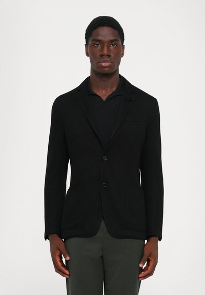 Mann in einem schwarzen strukturierten Blazer über einem schwarzen Hemd mit dunkelgrauen Hosen, stehend vor einem schlichten hellen Hintergrund.