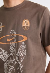 Brun bomulds t-shirt med hvid pixeliseret grafik af en figur med vinger, suppleret med orange logoer og cirkulære accenter.