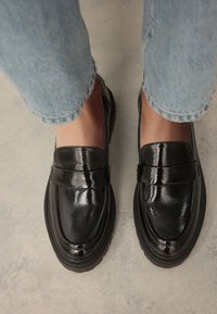 Black patent leather loafers with a slim profile and decorative strap, featuring a thick, textured sole. Worn with cuffed light blue jeans.