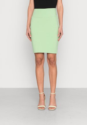 Femme portant une jupe crayon vert clair arrivant au genou, un haut noir et des sandales à talons hauts vert pâle, debout devant un fond uni.