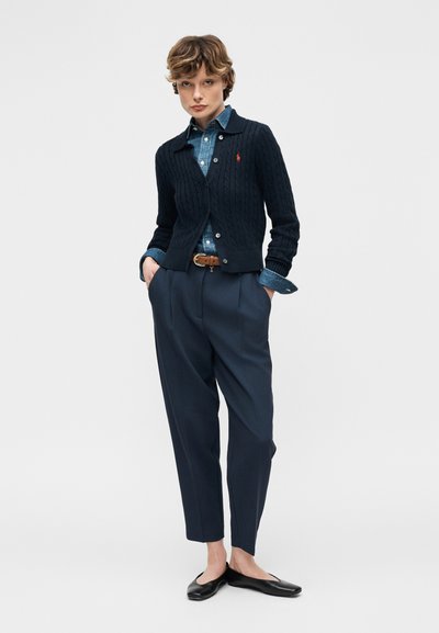 Γυναίκα που φοράει μπλε ζακέτα με πλεκτά σχέδια πάνω από μπλε πουκάμισο τζιν, μπλε πλισέ παντελόνια, καφέ ζώνη και μαύρες μπαρέτες, στέκεται με τα χέρια στις τσέπες.