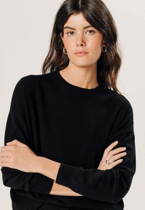 Vrouw met lang donker haar, gekleed in een zwarte trui en zilveren oorringen in de vorm van hoepels, staand met gekruiste armen tegen een effen lichte achtergrond.