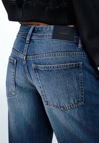 Traperice u srednje plavoj boji, s klasičnim dizajnom s pet džepova, vidljivim šavovima i crnom etiketom na pojasu.