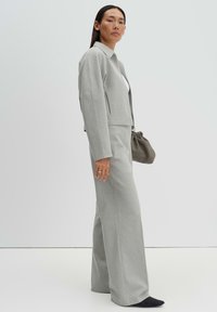 Donna con capelli lunghi e scuri, che indossa un completo grigio chiaro con pantaloni a gamba larga, top bianco, scarpe nere e tiene una piccola borsa grigia.