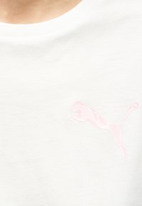 Λευκό πουκάμισο με ένα μικρό, ανοιχτό ροζ λογότυπο γάτας που πηδά στο στήθος, φορεμένο από ένα άτομο με εμφανή περιοχή λαιμού και ώμου.