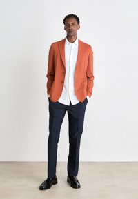 Orange strukturerad kavaj, vit skjorta och mörkblå byxor. Svarta skor fulländar outfiten och visar på en skräddarsydd passform och modern design.