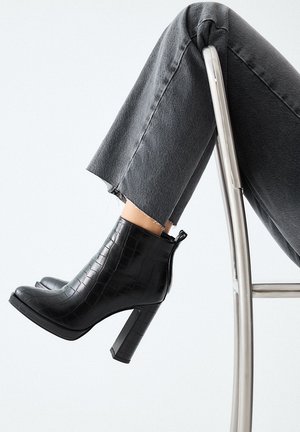 Botas de tobillo negras con estampado de cocodrilo, con puntera cuadrada y tacón alto. Combinadas con unos jeans grises deshilachados, descansando sobre una silla de metal.