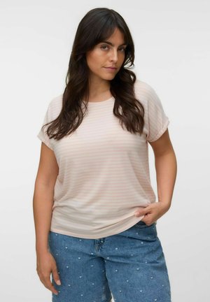 Kvinde med langt mørkt hår iført en lyserød stribet T-shirt og blå jeans med små hjerte mønstre, står mod en ensfarvet baggrund.