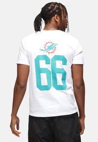 Camiseta blanca de manga corta con el número "66" en color azul verdoso en la parte posterior y un logo de delfín encima, mostrando una textura de algodón suave.