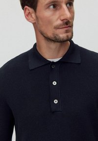 Tummansininen neulepolo, jossa on litteä kaulus, kolme napinläpeä, teksturoitu kangas ja erottuvat napit. Tiukka istuvuus.