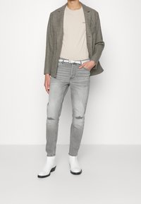 Persoon in een geblokte blazer, beige t-shirt, lichtgrijze ripped jeans met een witte riem, en witte enkellaarsjes staat tegen een witte achtergrond.