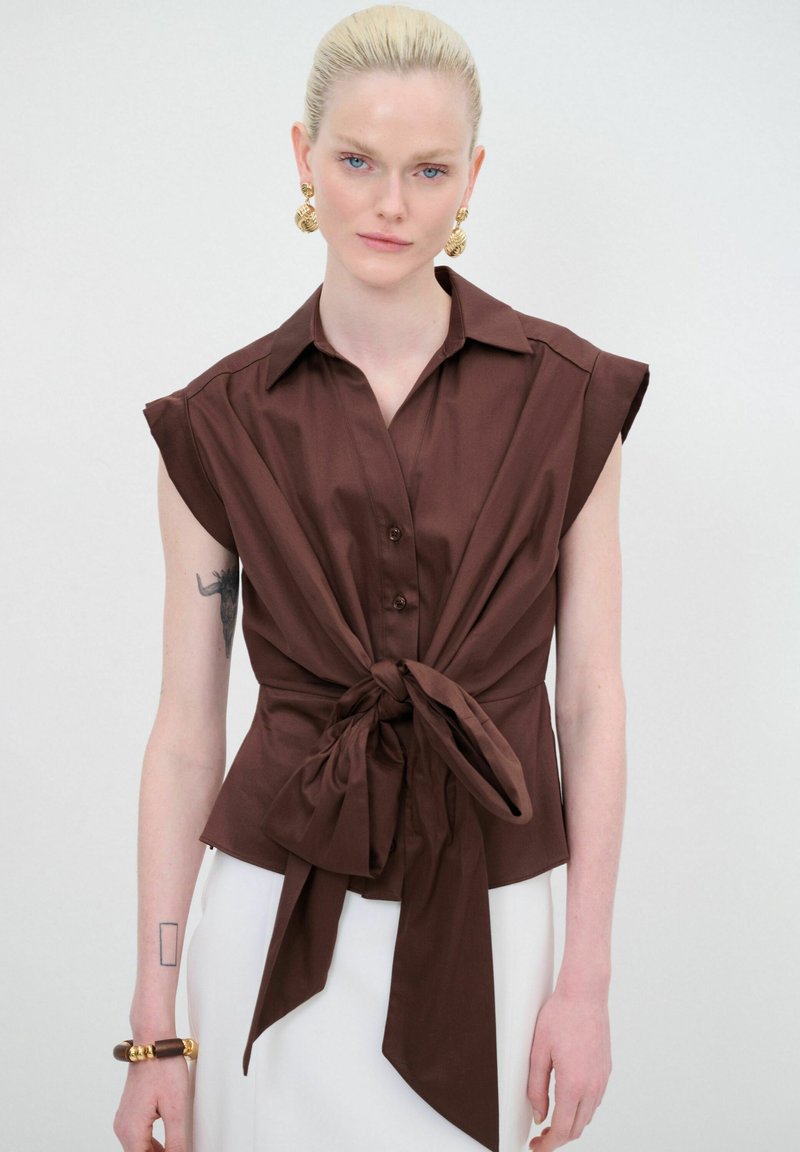 Blusa marrone a maniche corte con chiusura a bottoni e dettaglio anteriore legato. Tessuto morbido con una forma strutturata e design del colletto classico.