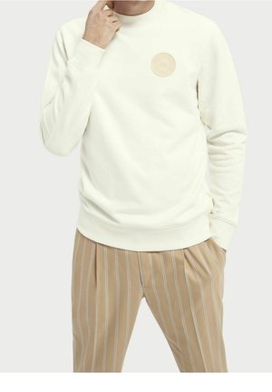 Mann trägt einen elfenbeinfarbenen Pullover mit rundem Aufnäher auf der Brust und beigefarbene, fein gestreifte Hose, Hand nahe am Gesicht, vor einfachem Hintergrund.